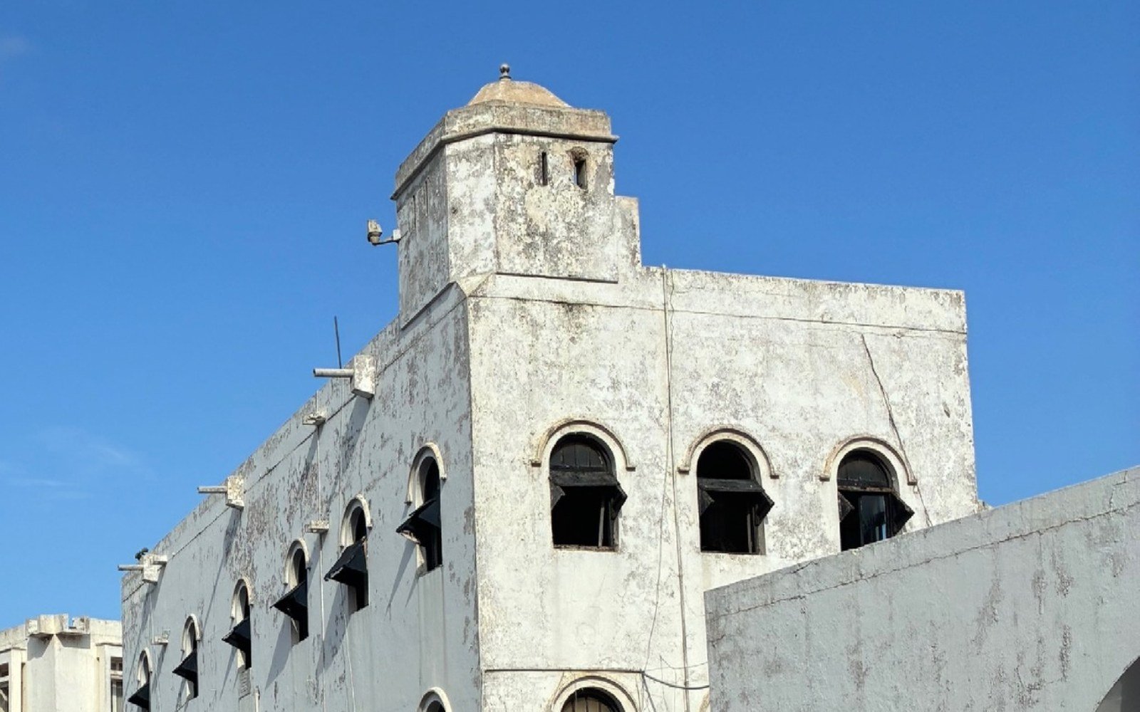 A white-washed historic stone building with a square corner tower and rows of arched windows against a clear blue sky