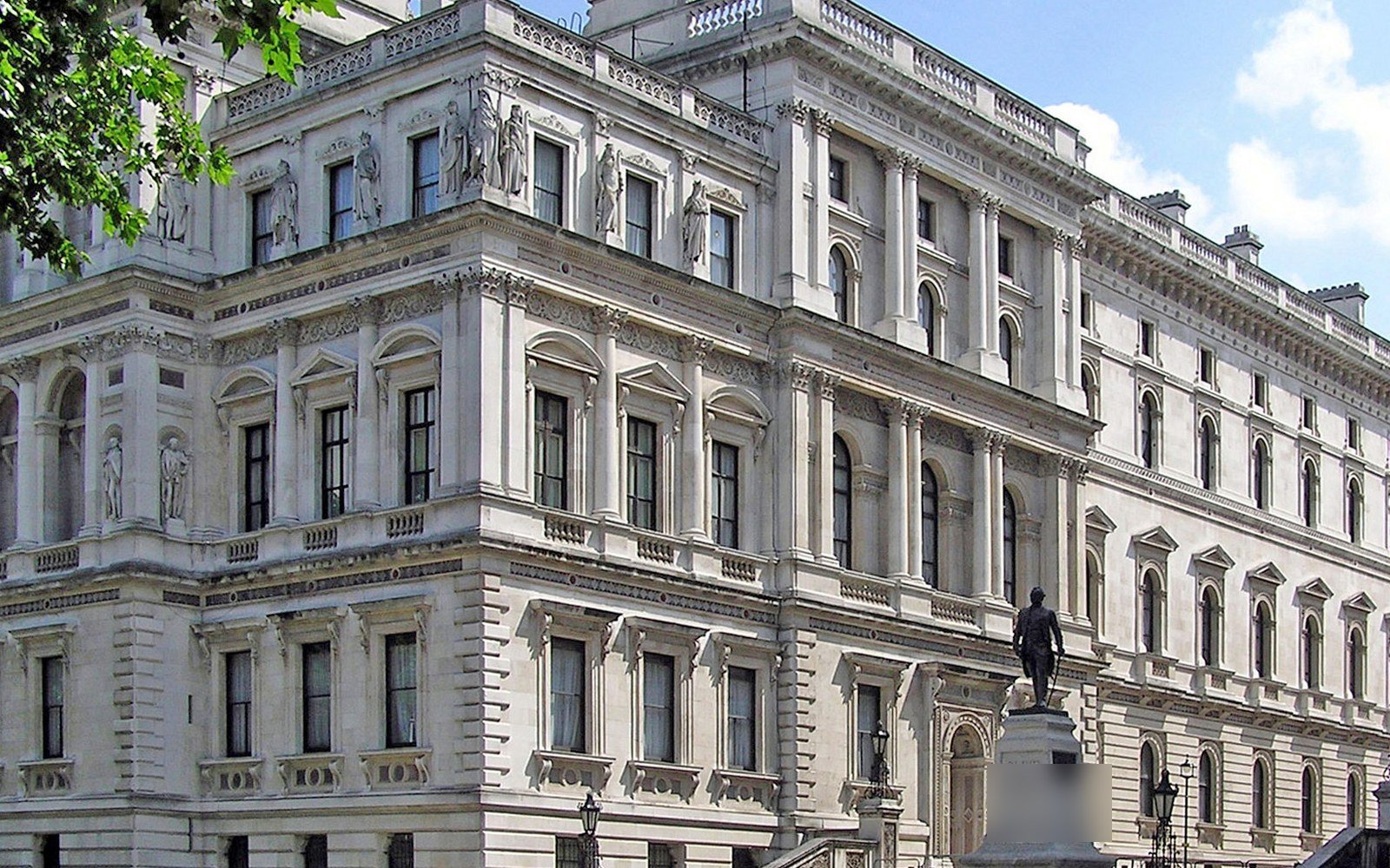 The ornate stone facade of a large Victorian government building with rows of tall windows, a statue on a plinth and a tree in the corner