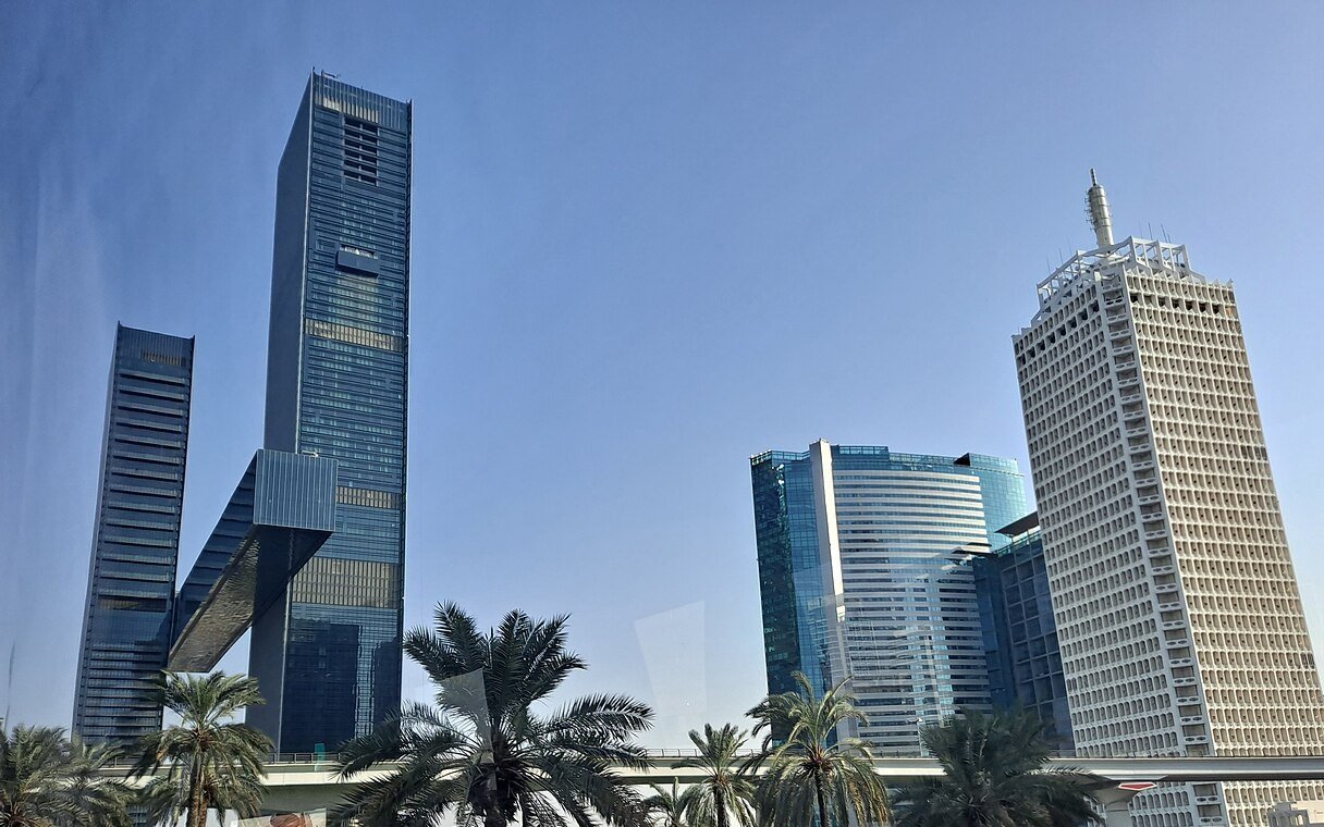 Office towers beside Sheikh Zayed Road in Dubai under a clear sky, with palm trees and an elevated rail line in front