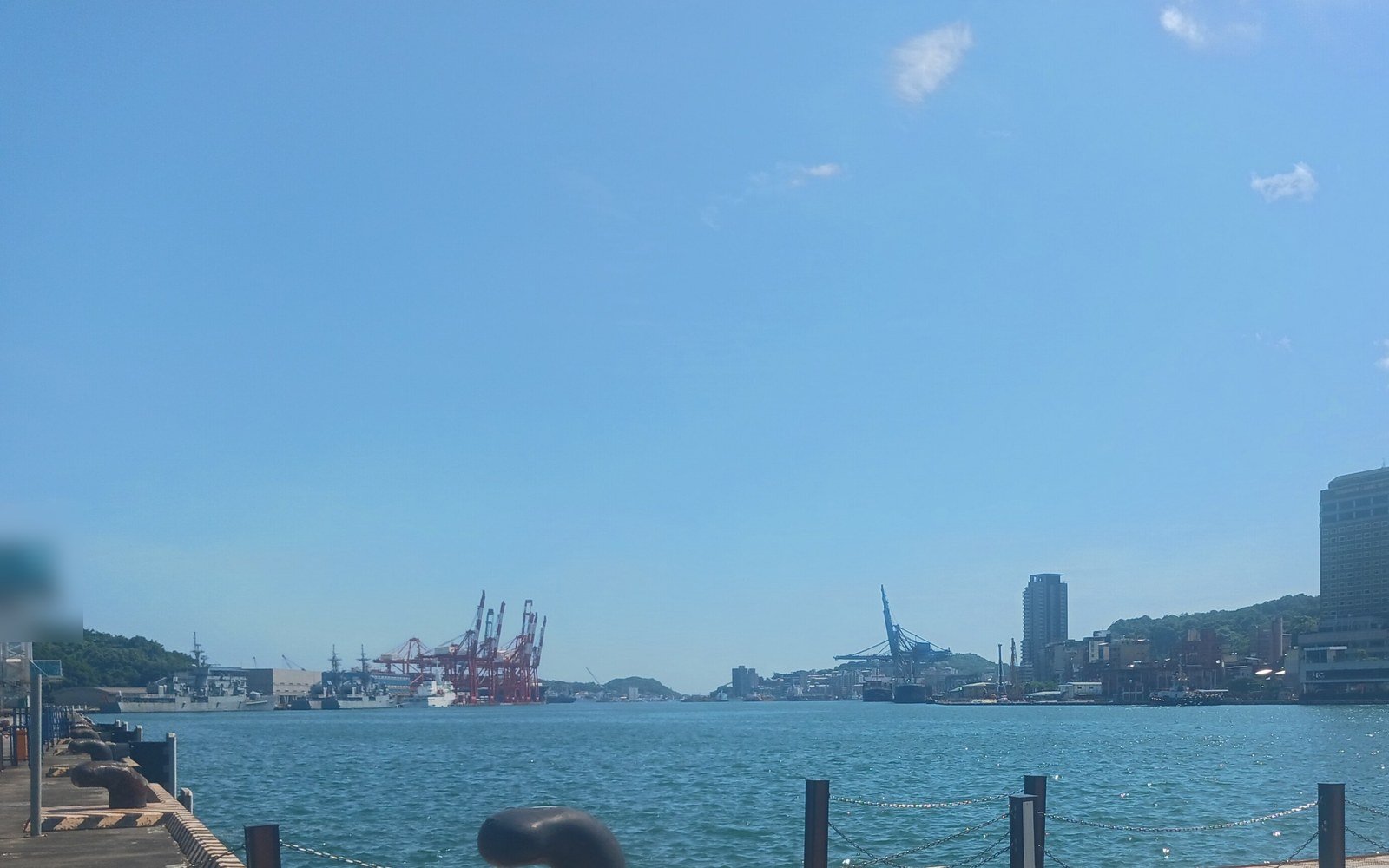 A calm blue harbour seen from a quayside with mooring bollards, with red container cranes and a hillside town on the far shore under a clear sky