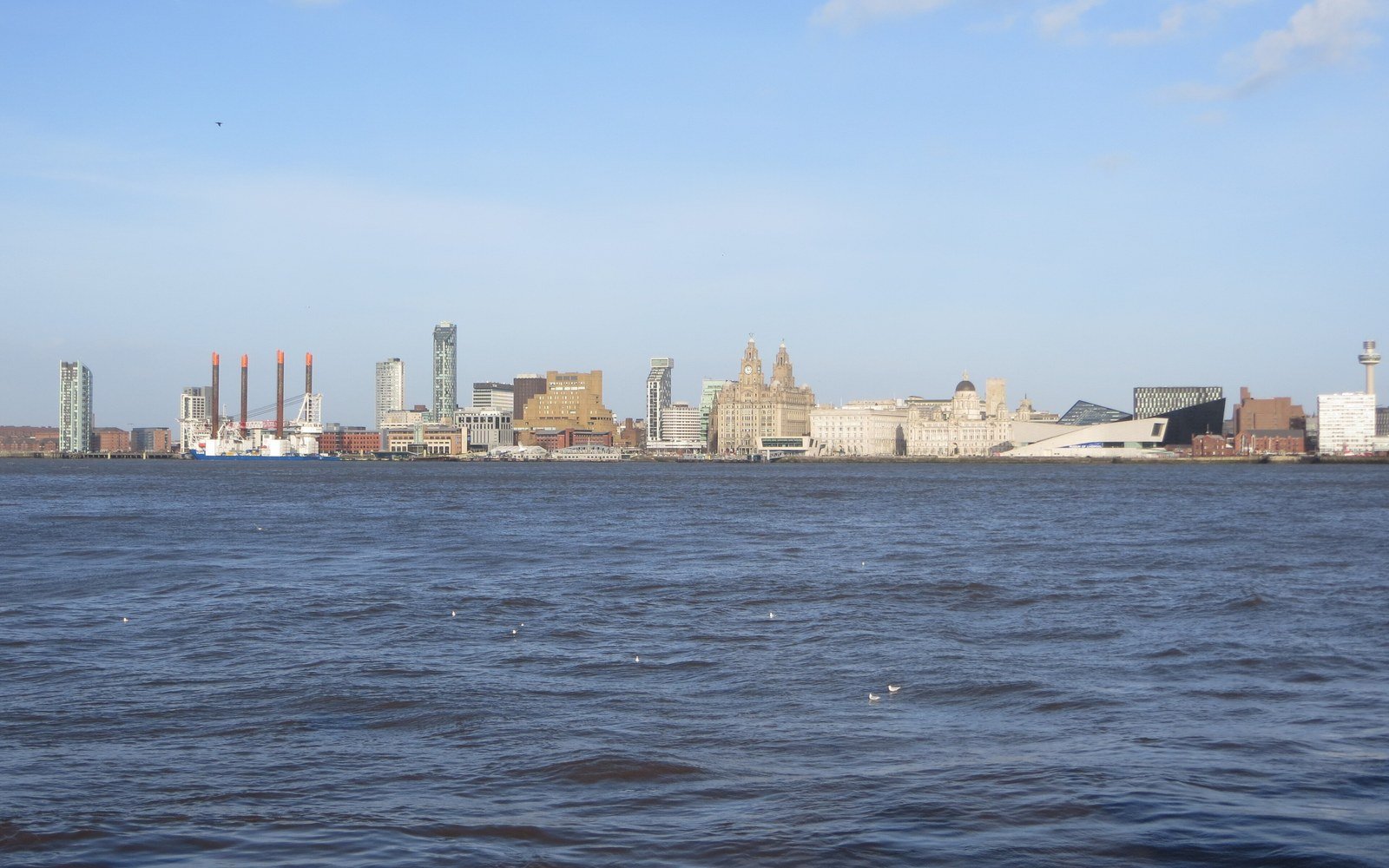A city waterfront of towers, domed historic buildings and chimneys seen across a wide choppy river under a blue sky