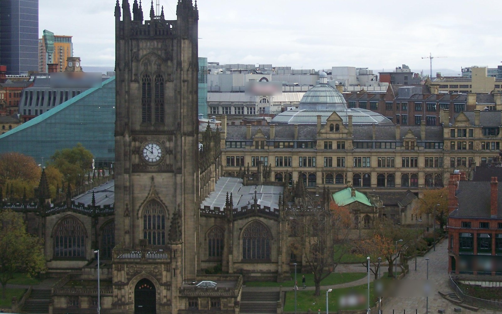 A Gothic cathedral with a clock tower in front of Victorian commercial buildings with a glass dome, a sloping glass building and city roofs beyond
