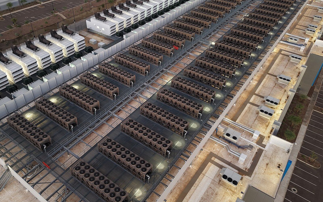 Rows of rooftop cooling units on a large data centre, with generator enclosures alongside, seen from above at dusk