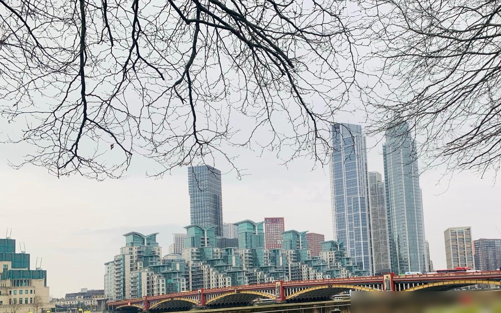 Tall glass residential towers and stepped apartment blocks beyond a red and gold arched bridge over a river, framed by bare tree branches