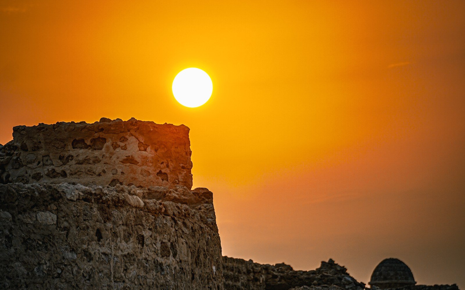 The sun sits low over a weathered stone bastion at Bahrain Fort against an orange sky, with a small stone dome standing beyond the wall