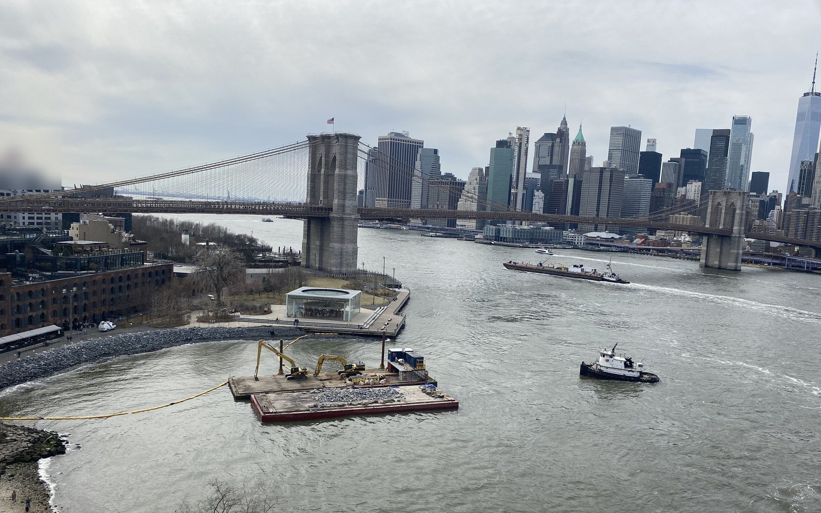 A wide grey river with a tug and a work barge in the foreground, a suspension bridge crossing it and a cluster of tall city towers beyond