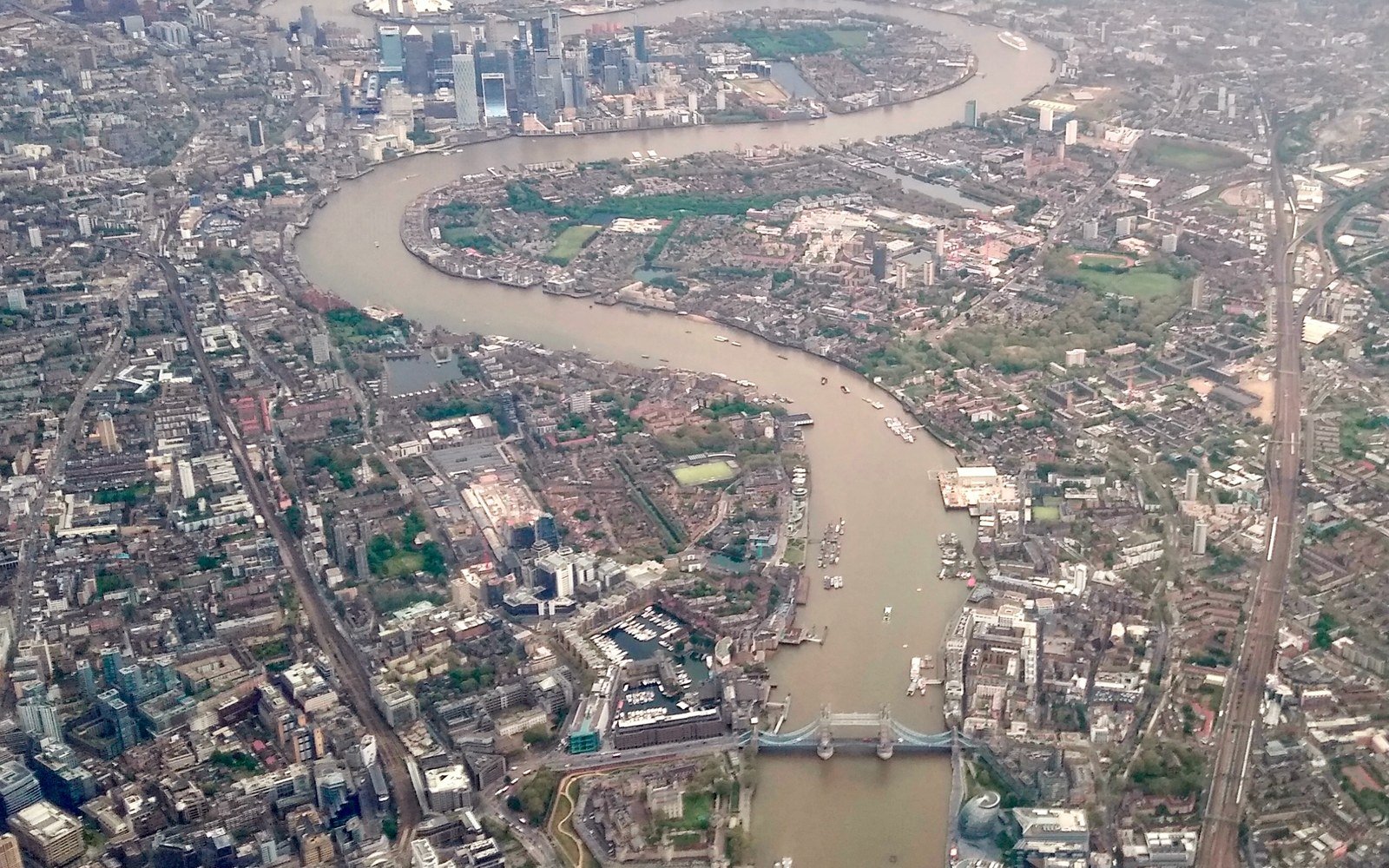 An aerial view of a wide brown river curving through a dense city, with a bascule bridge in the foreground and a cluster of tall towers in the distance