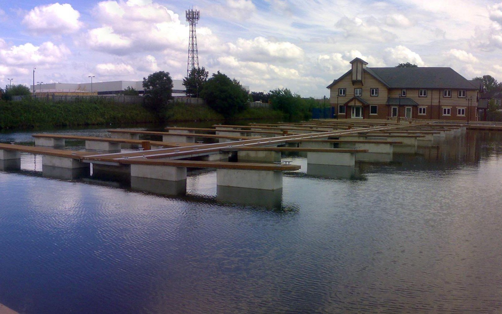A still canal basin with rows of concrete mooring pontoons, a lattice mast and a brick office building beyond under a cloudy sky