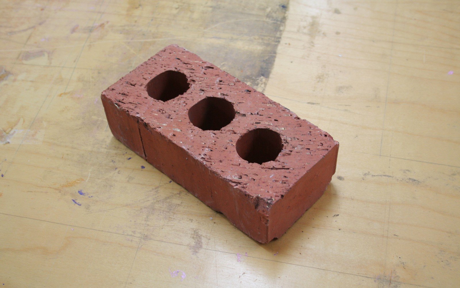 A single perforated red clay brick of the kind used in building work, lying on a plywood workbench