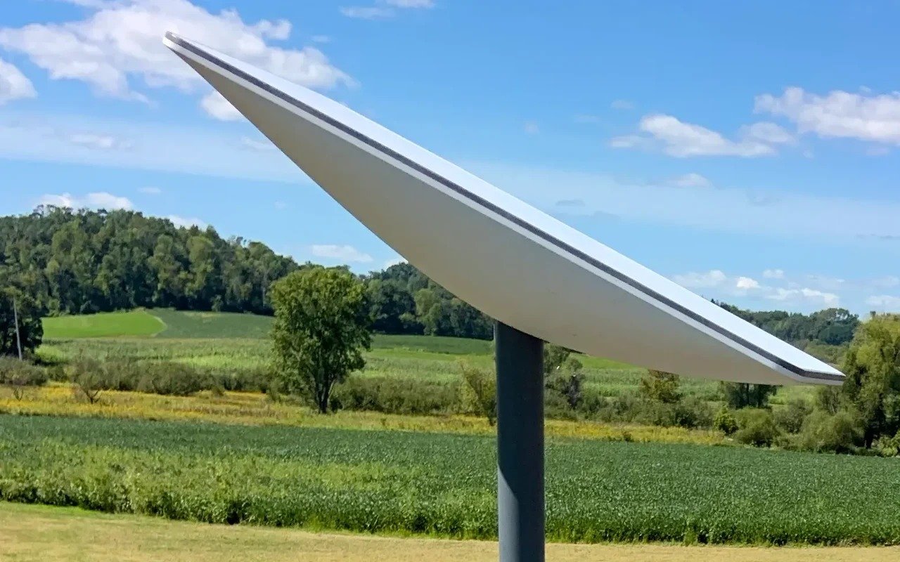 A flat white Starlink internet antenna on a grey pole, tilted towards the sky above green farmland and woods