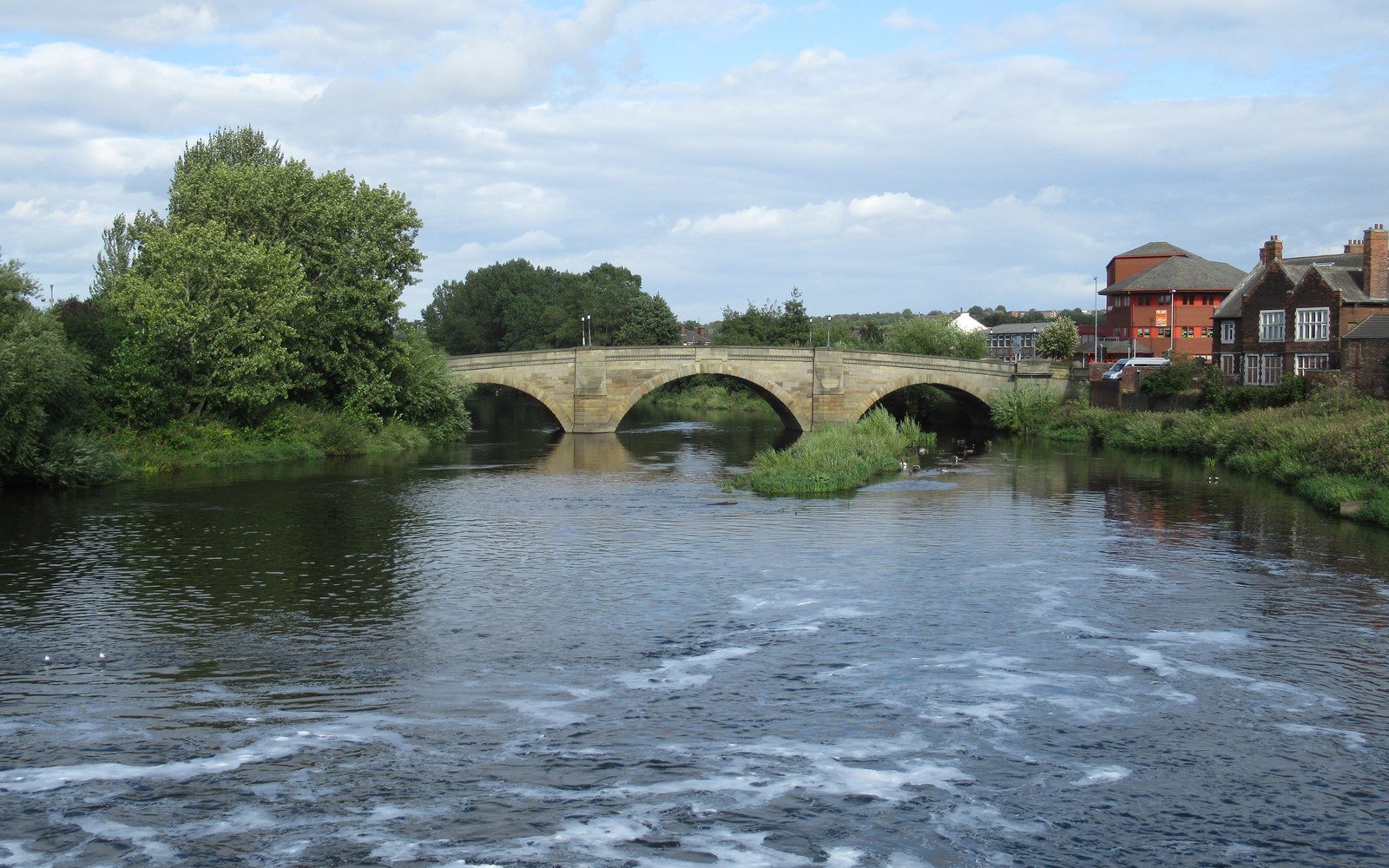 A three-arched stone road bridge over a wide river with fast shallow water in the foreground, trees on the left bank and brick houses on the right