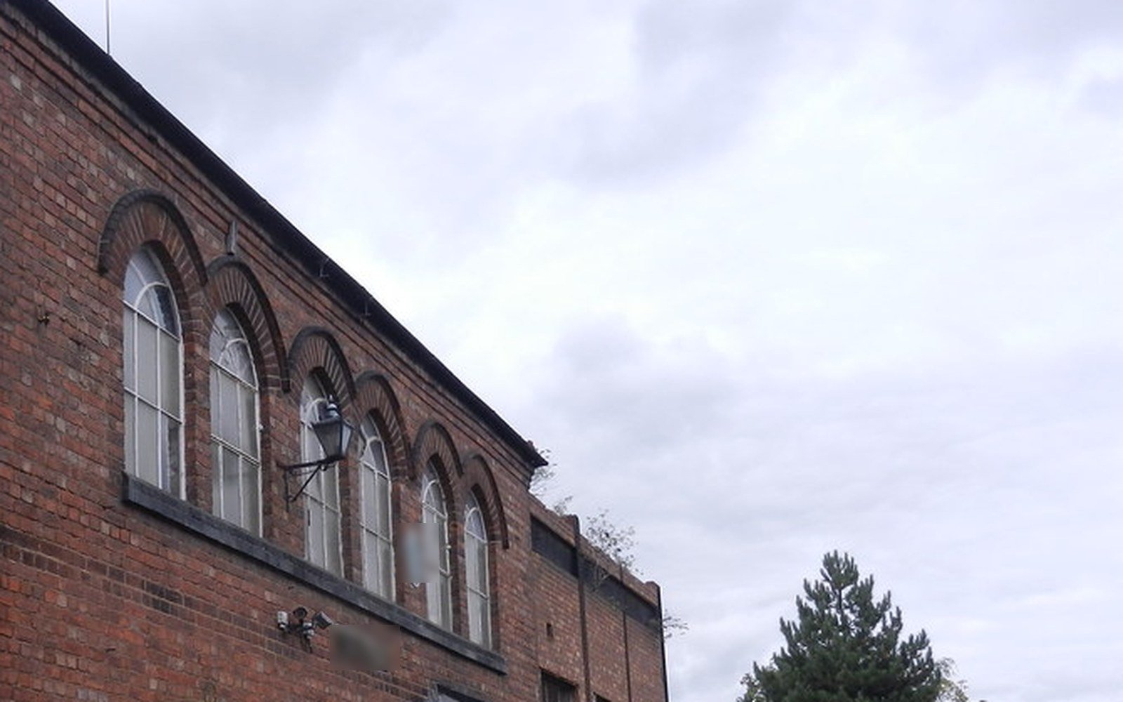The upper storey of an old red brick building with a row of tall arched windows and a wall lamp, under a cloudy sky, with a pine tree beyond