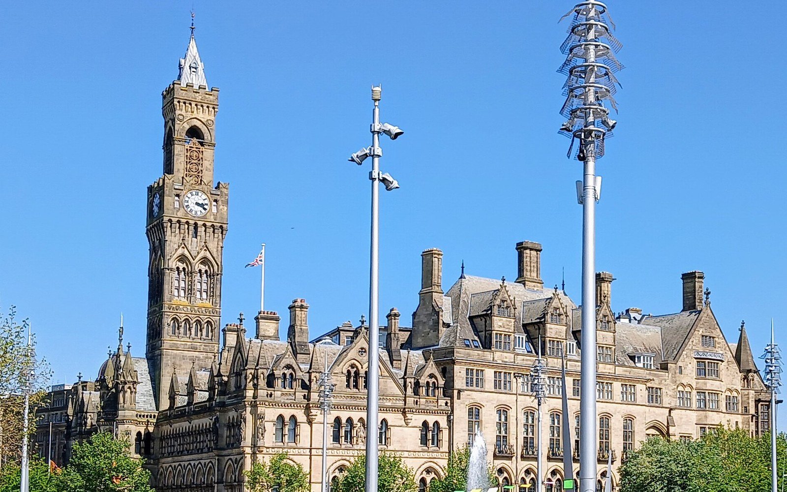 A Victorian Gothic town hall with a tall clock tower and stone facade under a clear blue sky, with tall steel lighting masts and trees in front