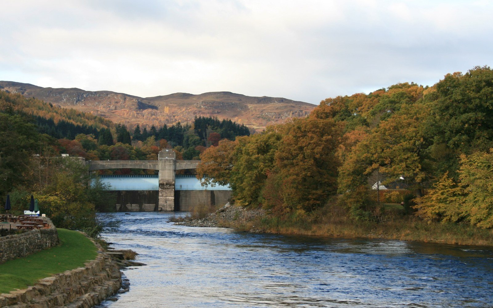 A fast river flowing towards a concrete dam between banks of autumn trees, with brown hills rising behind under a pale sky