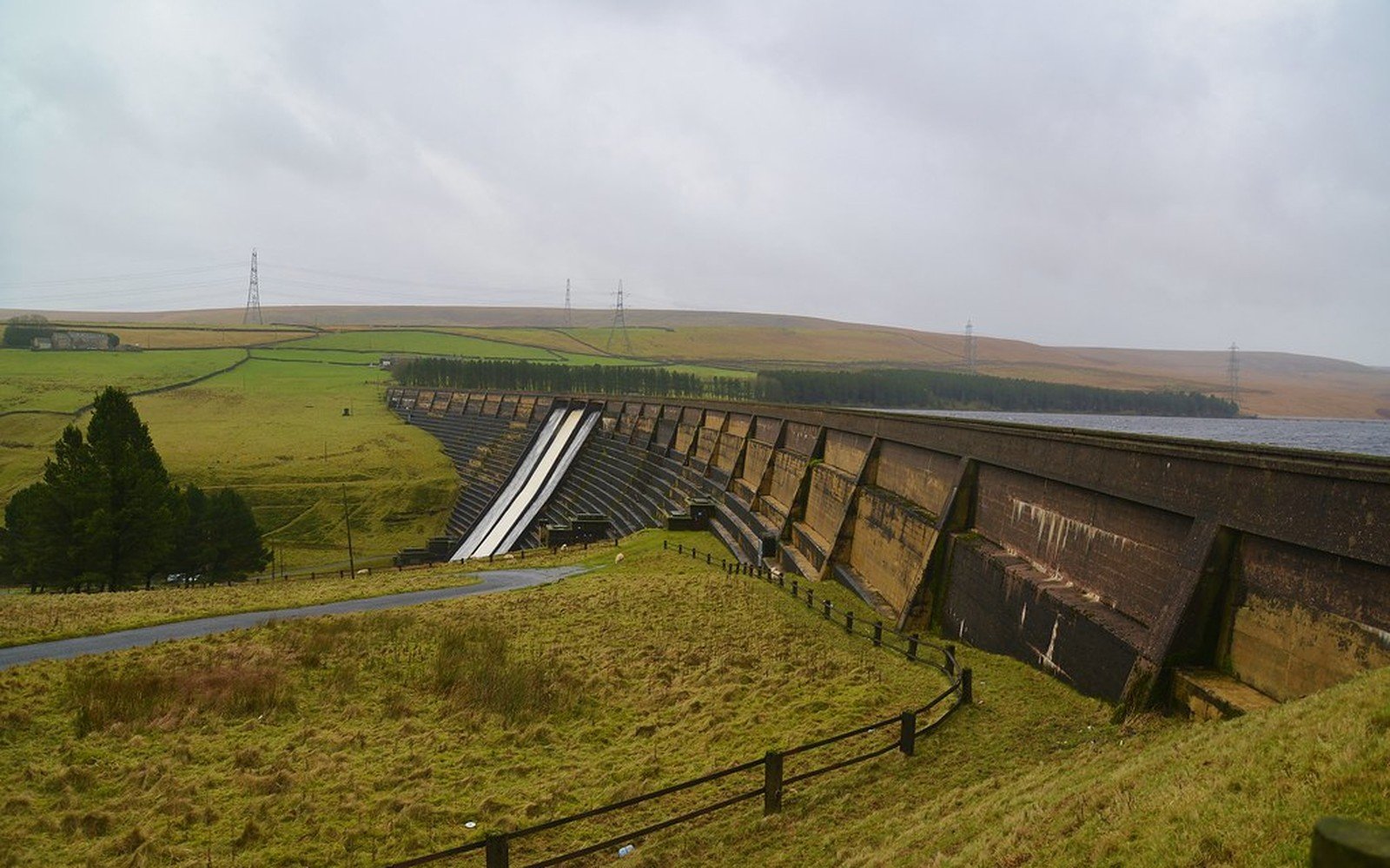 A long stone dam with a spillway curving across a green valley, with moorland, pylons and an overcast sky beyond